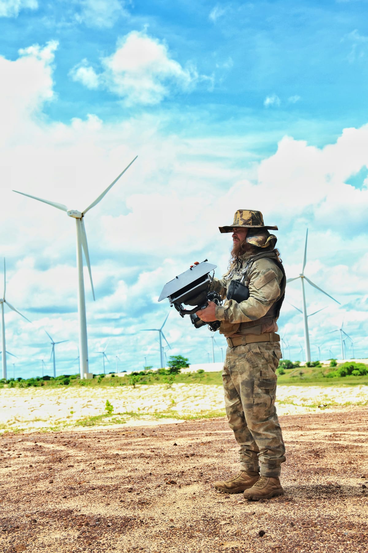 A drone operator in camouflage conducting a pre-flight inspection at a wind farm with turbines in the background.