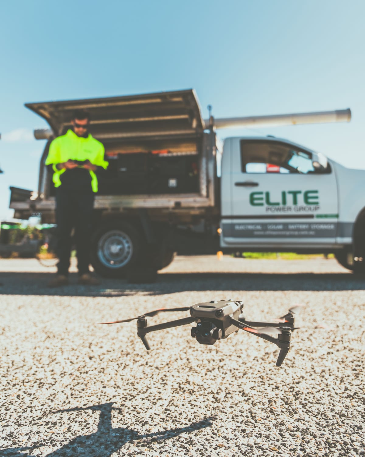 Commercial DJI Mavic 3 drone hovering above a gravel job site in front of a white service truck with a worker in a high-visibility jacket operating the remote controller.