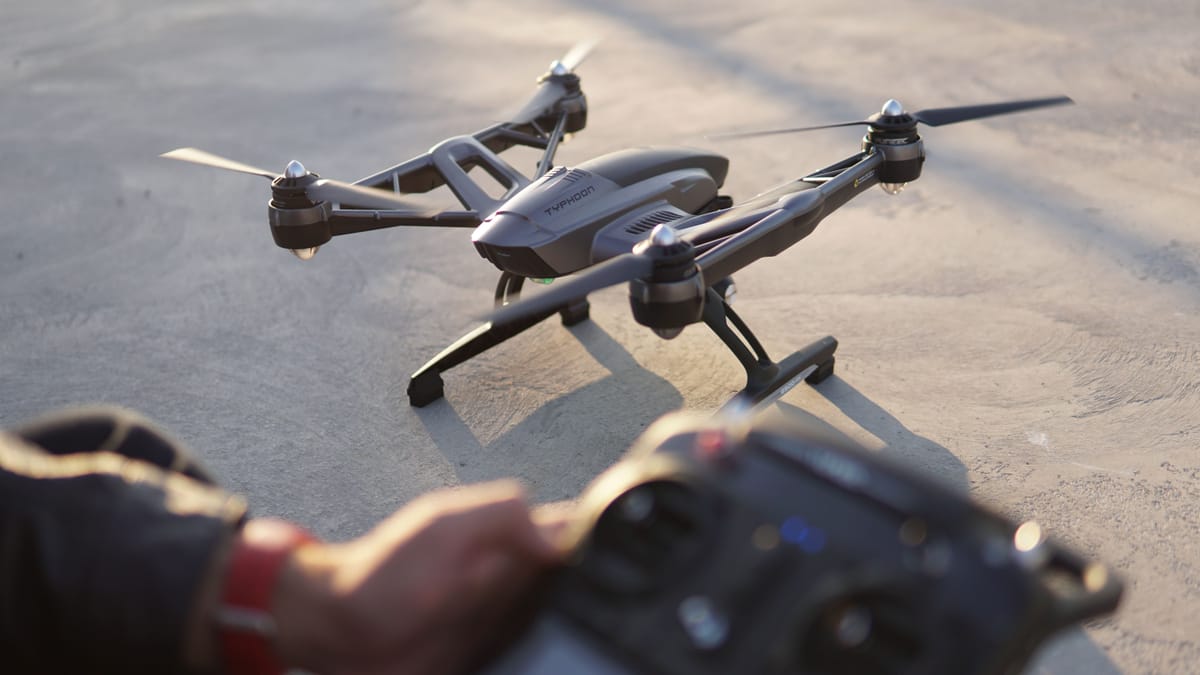 Top-down view of a quadcopter on pavement beside a drone operator holding a remote controller.