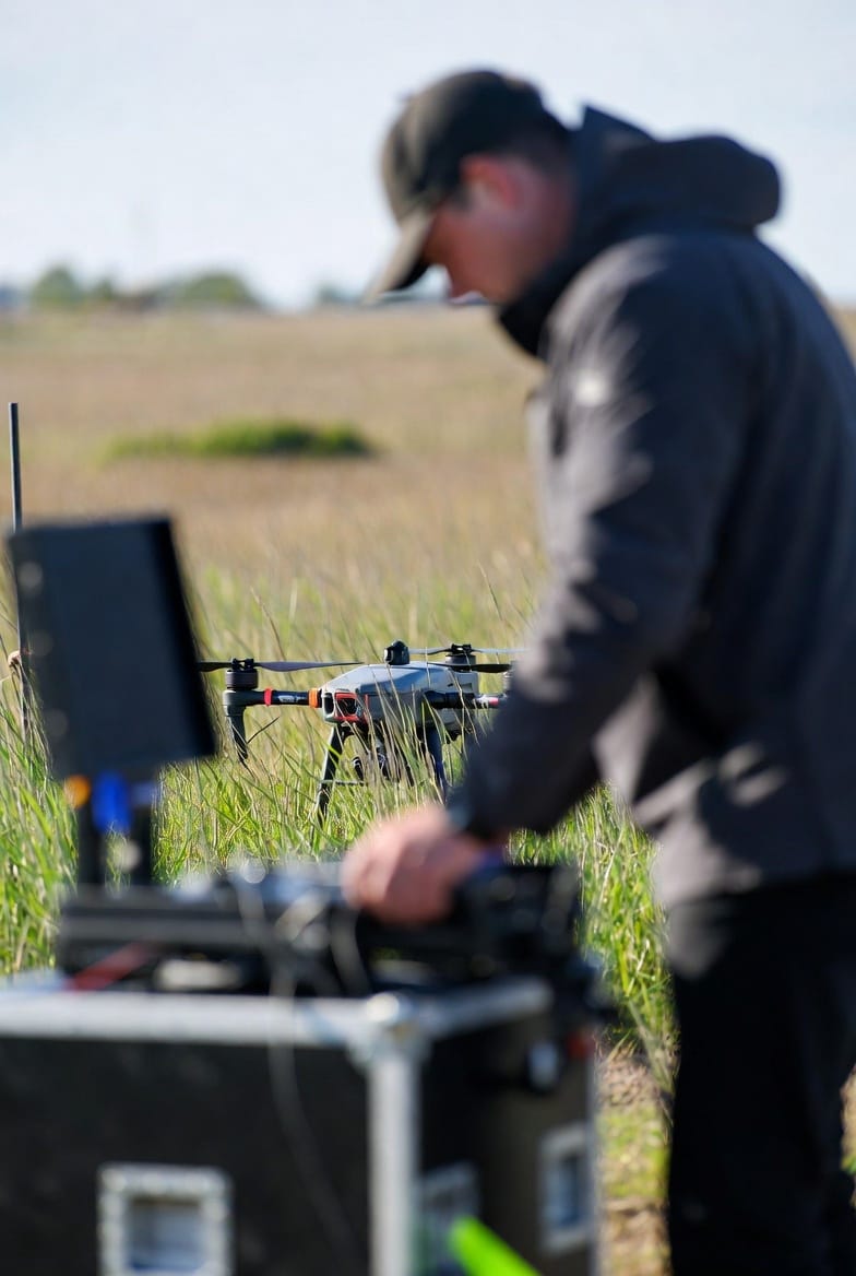 Drone operator checking equipment outdoors with drone visible in background.