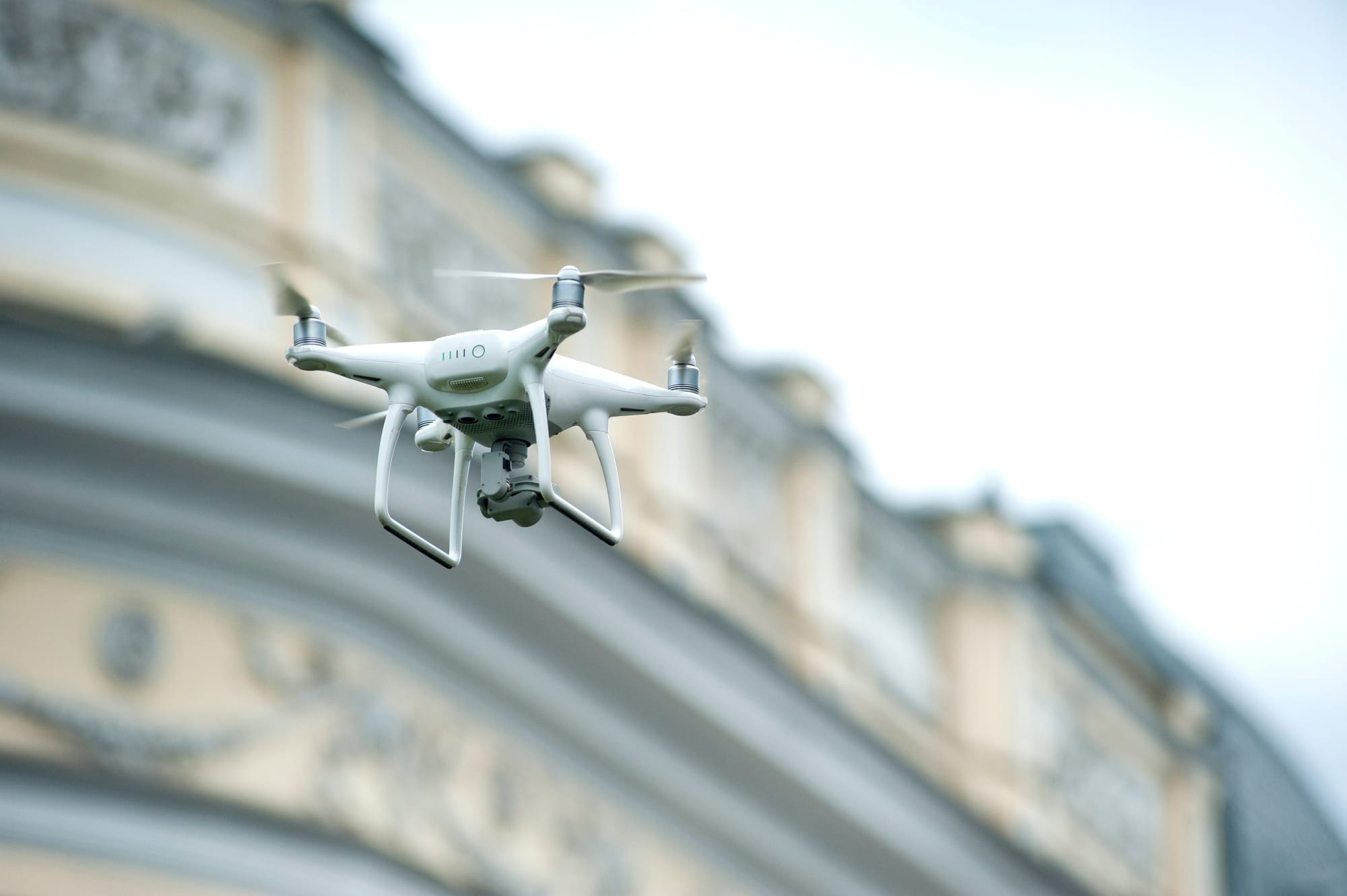 White quadcopter drone hovering against a blue sky near a domed urban building.