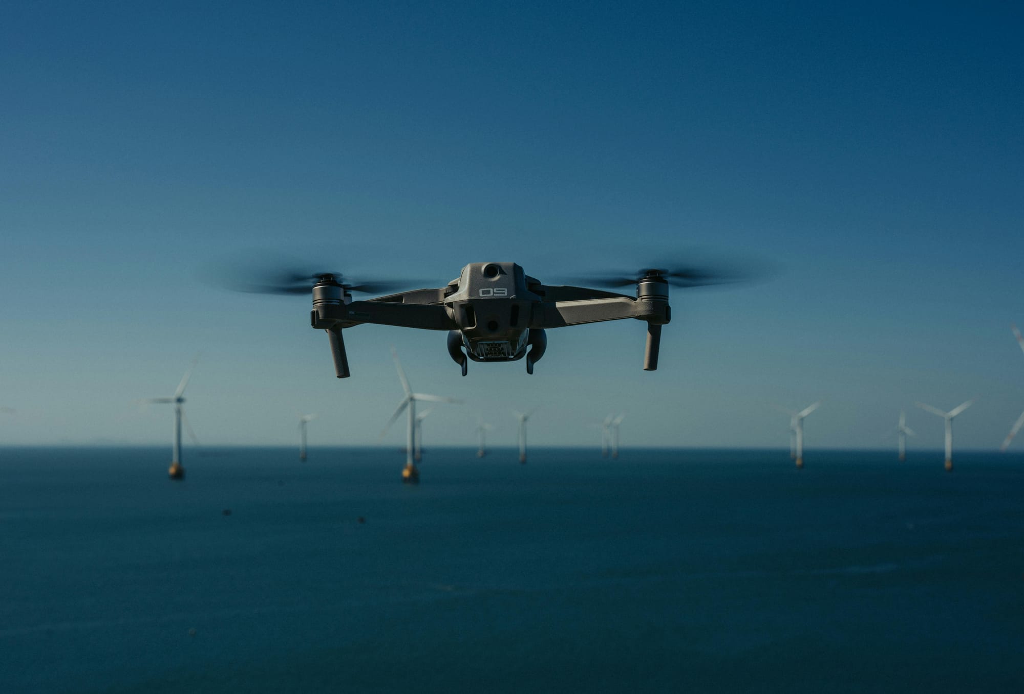 Drone in flight over ocean with offshore wind turbines visible in the background.