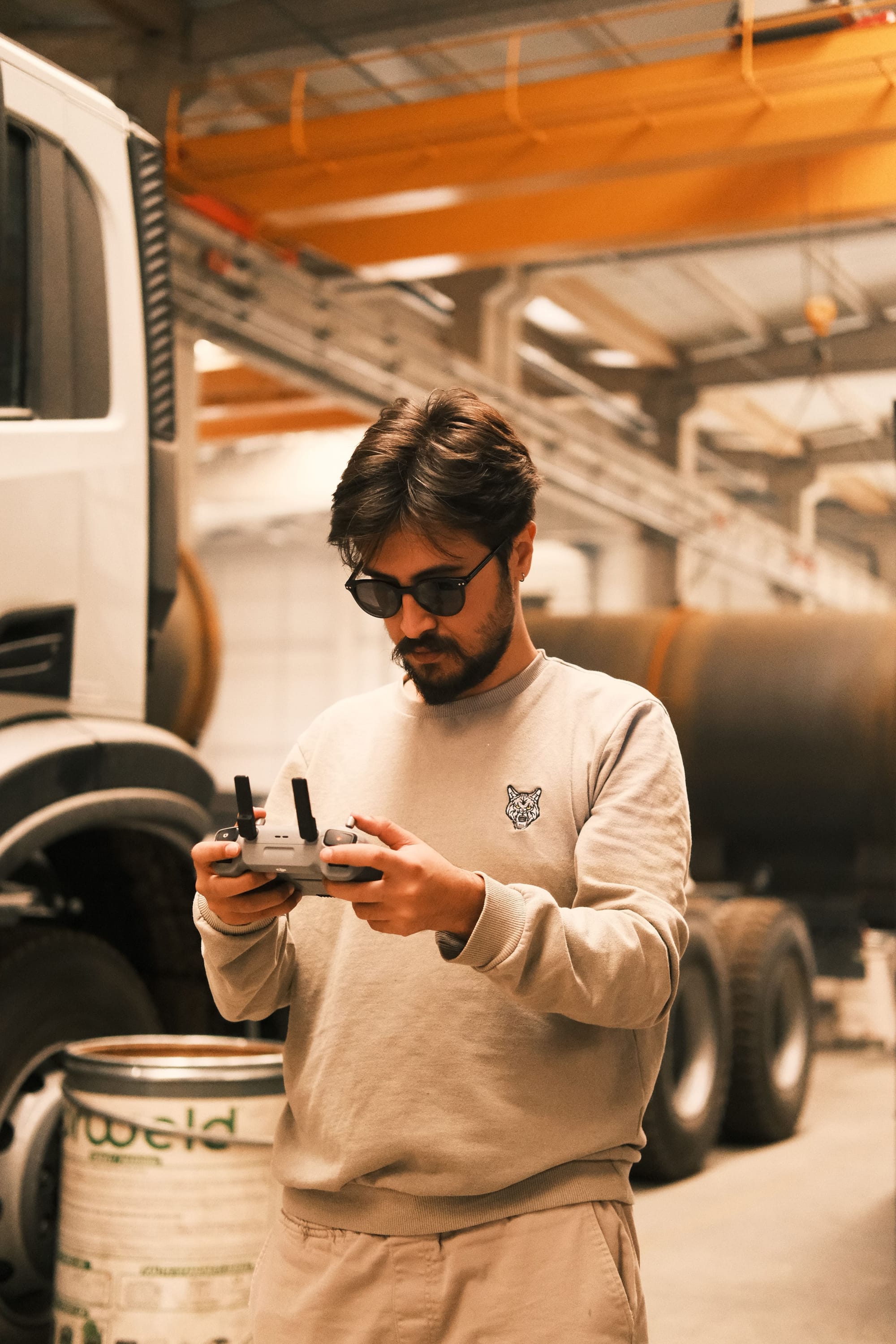 Drone operator using a remote controller inside an industrial workspace with heavy equipment in the background.