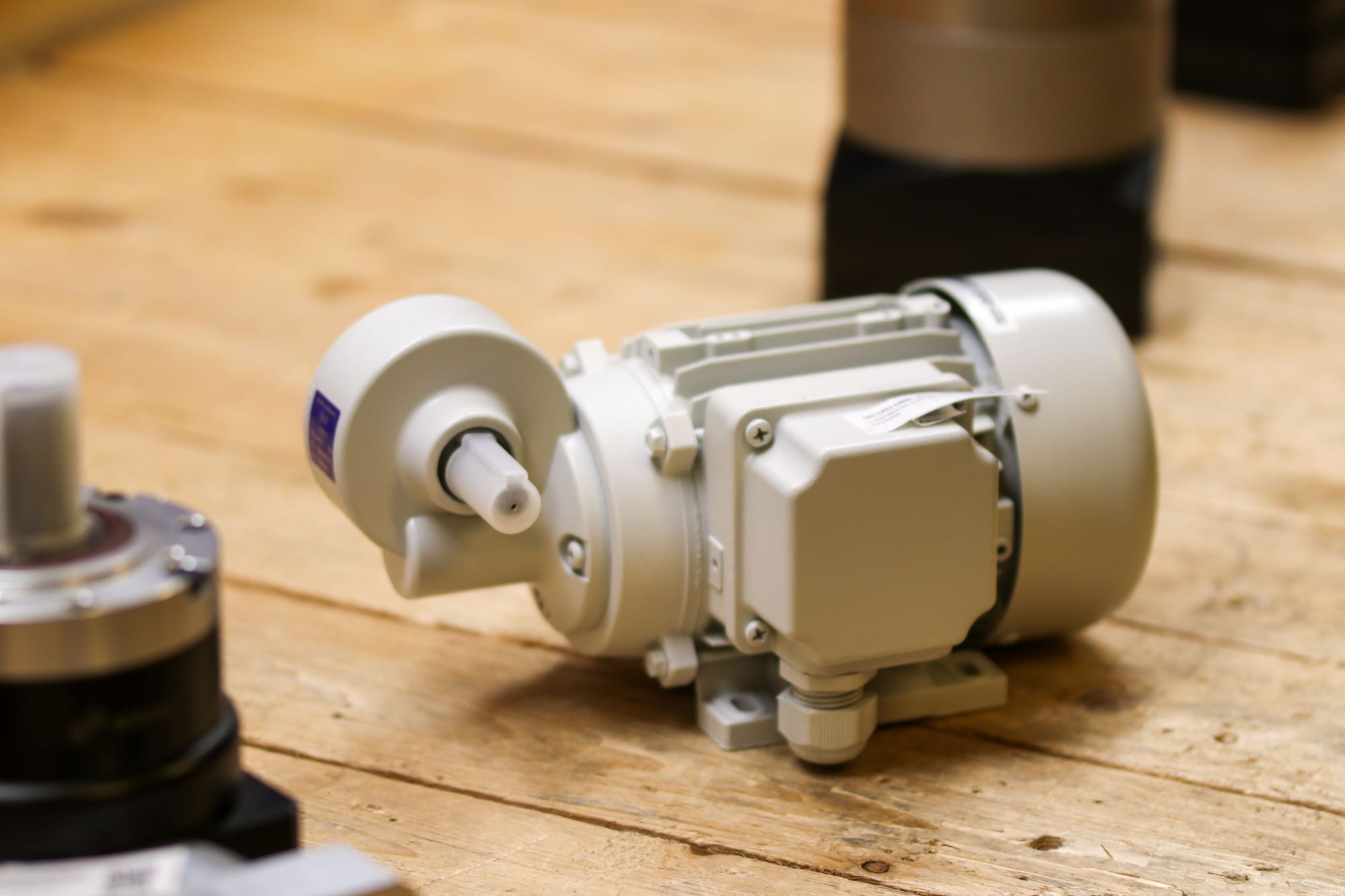 White industrial electric motor with output shaft and terminal box sitting on a wooden workshop surface next to a black cylindrical component.