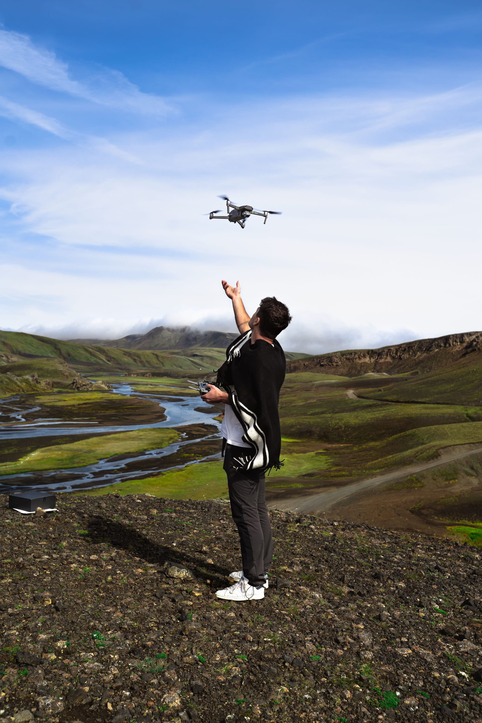 Drone operator standing on a green hillside and flying a quadcopter above a winding river landscape.