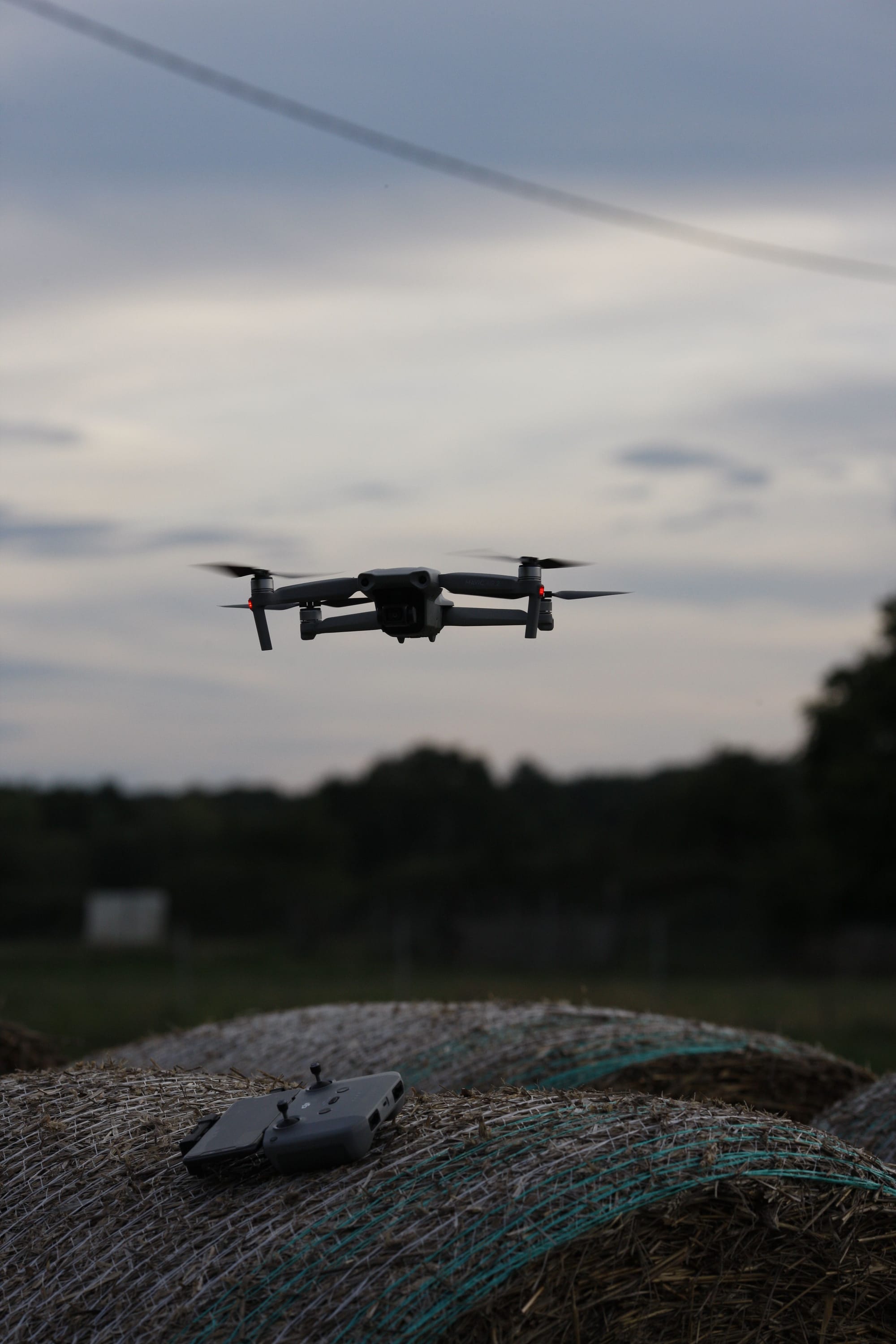 Drone hovering above a remote controller in a rural field at dusk.